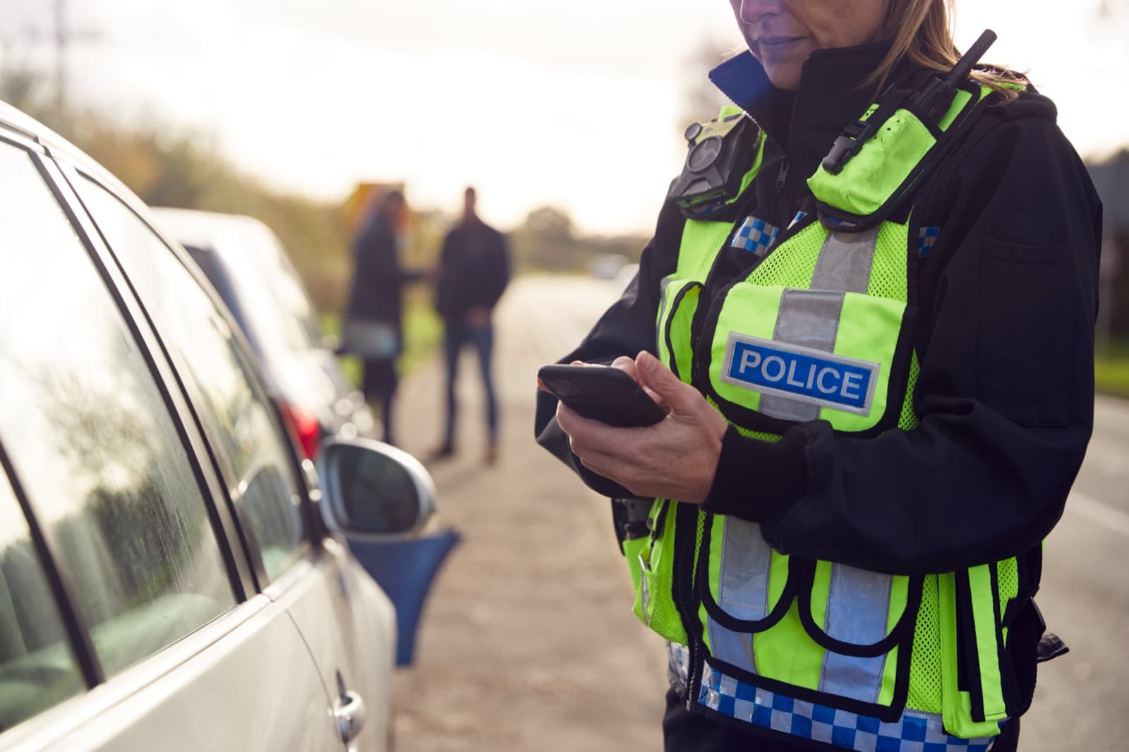 An officer in a high visibility vest making notes on a phone beside a stopped car, with two figures further up the road