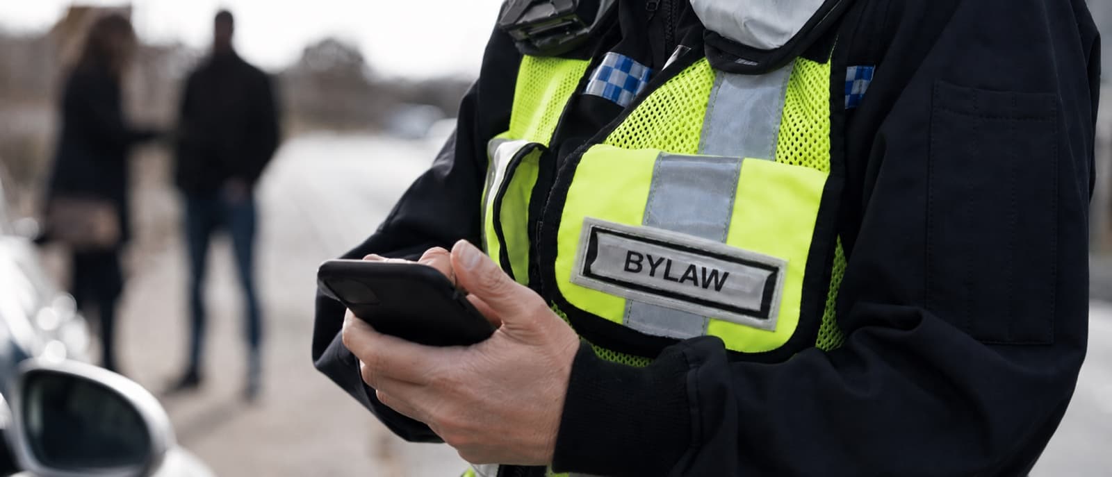 A bylaw officer in a high visibility vest making notes on a phone at the roadside