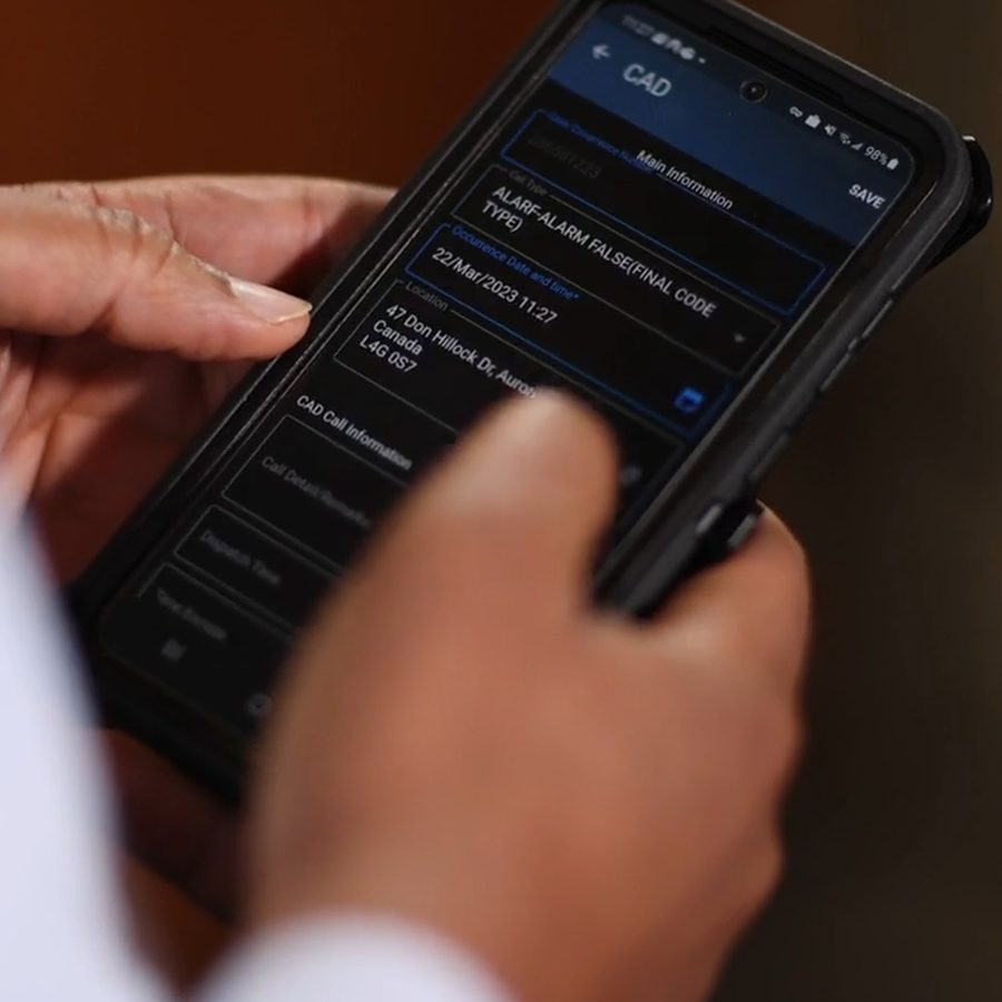 An officer's hands on a ruggedized handheld running IMP eNotes, the CAD screen showing a call type, timestamp and York Region address carried through automatically